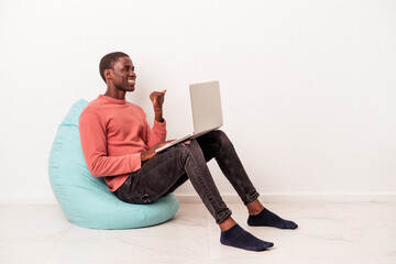 Young African American man sitting on a puff using laptop isolated on white background points with...