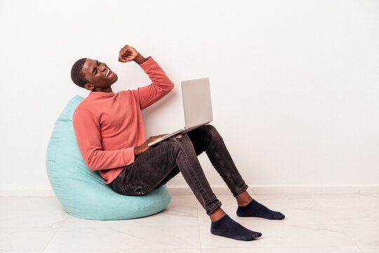 Young African American Man Sitting On A Puff Using Laptop Isolated On White Background Raising Fist After A Victory, Winner Concept.