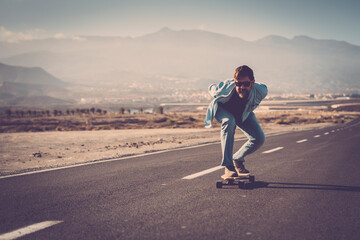Mature man in sunglasses with hands back riding his longboard on country road with mountains in background. Man skating or longboarding on mountain road