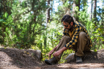 Fototapeta premium Mature male hiker in sunglasses tying shoelaces and getting ready for trekking in forest while sitting on rock. Man tying hiking shoelaces in forest before a long hiking tour in nature