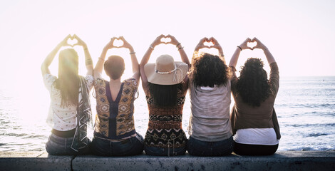 Rear view of five women sitting in a row on sea wall gesturing love by making heart shape symbol...