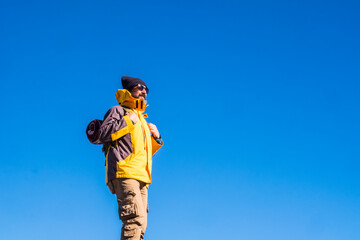 Mature man in backpack and sunglasses against blue sky. Male hiker in knit hat, sunglasses, backpack and yellow jacket admiring something interesting against blue sky