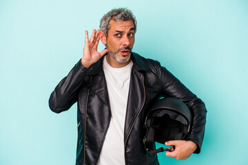 Middle age biker caucasian man holding helmet isolated on blue background  trying to listening a...