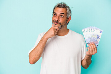 Middle age caucasian man holding bills isolated on blue background  looking sideways with doubtful and skeptical expression. - obrazy, fototapety, plakaty