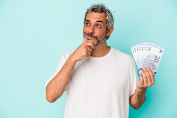 Middle age caucasian man holding bills isolated on blue background  looking sideways with doubtful...