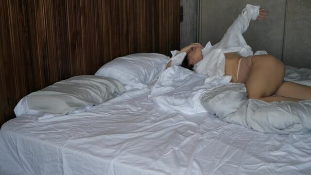 a young attractive woman in a white shirt and bare legs lies on a white bed in a hotel room and pulls her hands up. High quality FullHD footage