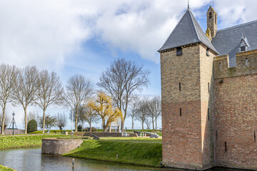 Obraz premium Bare trees, moat, green grass, Radboud castle tower with brick walls and canons in the background, sunny day with a blue sky and white clouds in Medemblik, Noord-Holland, Netherlands