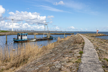 Fototapeta na wymiar Entrance to Oosterhaven, city harbor with its dike, gates and a small anchored boat, horizon, windmills and blue sky in the background, sunny day at Medemblik in Noord-Holland, Netherlands