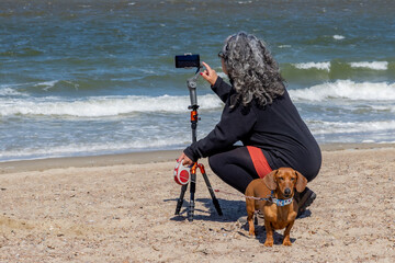Mature woman squatting with her back to camera on Renesse beach filming with her mobile phone...