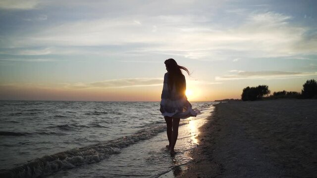 Young woman looking forward in dress walks along the wet sand on the seashore at sunset in summer