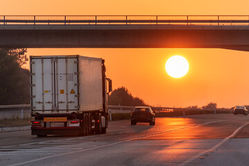 Container truck driving on the highway with the sun in front of it at sunset.