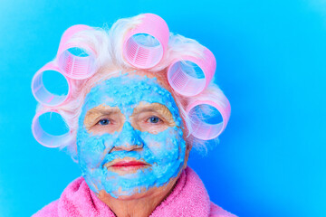 woman with blue cream moistening mask on face , puts curlers on white hair and wear pink bath robe in studio background