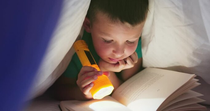 A young boy reading a book under the covers with a flashlight at dark night time