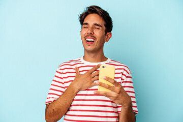 Young mixed race man holding mobile phone isolated on blue background laughs out loudly keeping hand on chest.