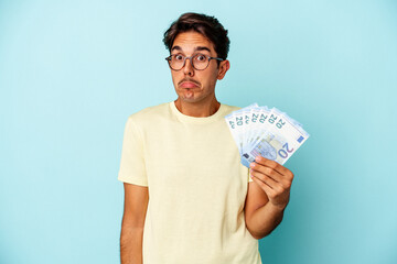 Young mixed race man holding bills isolated on blue background shrugs shoulders and open eyes confused.