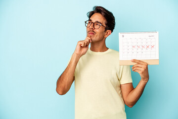 Young mixed race man holding calendar isolated on blue background looking sideways with doubtful...