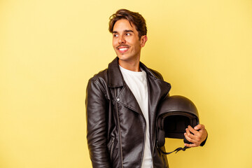 Young mixed race man holding helmet isolated on yellow background looks aside smiling, cheerful and pleasant.