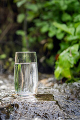 a glass of clean transparent drinking water in a transparent glass on a stone in a green forest near a stream or mountain spring. healthy food and diet, beautiful background. mountain stream closeup.
