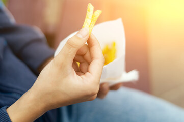Obraz na płótnie Canvas A young woman's hand takes out slices of yellow, appetizing French fries from a white paper bag. A young girl is eating delicious French fries right on the street, sitting on a bench in a city park.