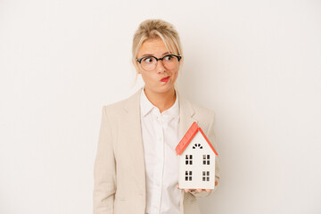 Young real estate agent woman holding a home model isolated on white background confused, feels...