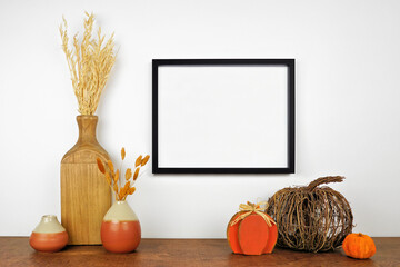 Mock up black frame hanging from a white wall with fall decor on a wood shelf. Autumn concept. Copy...