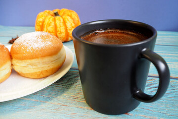 in a dark mug, hot natural coffee stands next to a white plate with a donut and a pumpkin on the back of a blue table. side view