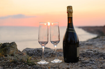 A bottle of champagne and two empty glasses stand on the mountain against the backdrop of a physical sunset. Selective focus on the bottle.
