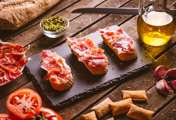 Iberian ham toasts on a slate plate on a brown wooden table with some ingredientes around it. Spanish typical food. Pork meat.