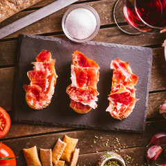 Iberian ham toasts on a slate plate on a brown wooden table with some ingredientes around it....