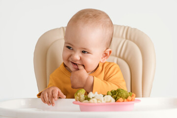 Funny baby on a feeding chair holds his fingers in his mouth. A plate of boiled vegetables in front of him