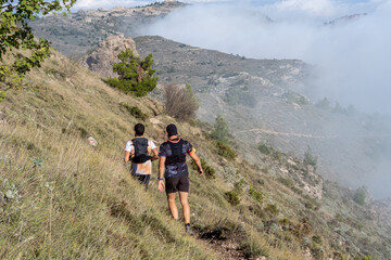 Obraz premium Two young men seen from behind running down a trail in the mountains, on a sunny day with fog. 
