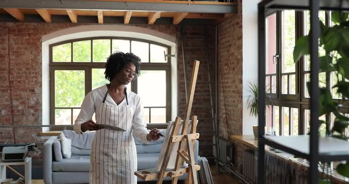 Fine-art, creative hobby concept. Young curly-haired African artist female painter standing in workshop holds palette and paintbrush, drawing a picture on canvas, smile feels inspiration enjoy process
