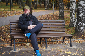 A man of European appearance sits on a bench in a park in autumn.