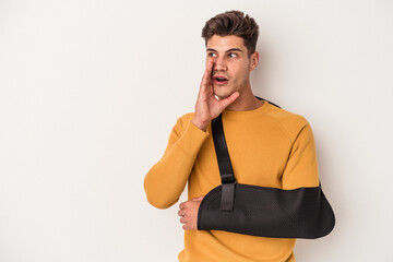 Young caucasian man with broken hand isolated on white background is saying a secret hot braking news and looking aside