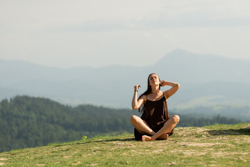 Beautiful brunette female sitting in pose of lotus in Carpathian  mountains and doing meditation, touching hair. Young woman in brown dress in practice of pranayama, enjoy nature, looking at camera.