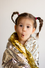 Portrait of pretty and emotional little girl with opened mouth looking at camera, covering gold paper for present. Kid wearing silver foil. Concept of New Year and Christmas holiday.