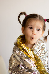 Portrait of pretty and emotional little girl with opened mouth looking at camera, covering gold paper for present. Kid wearing silver foil. Concept of New Year and Christmas holiday.