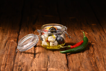 Feta cheese, pickled peppers and olives in a storage jar on the wooden table.