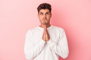 Young caucasian man isolated on pink background holding hands in pray near mouth, feels confident.