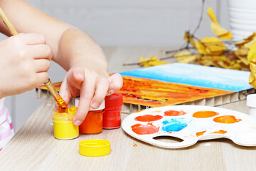 The child makes a craft or drawing. Children's hands dip a brush into yellow paint. In the background, autumn rowan leaves. There is a craft on the table - an application