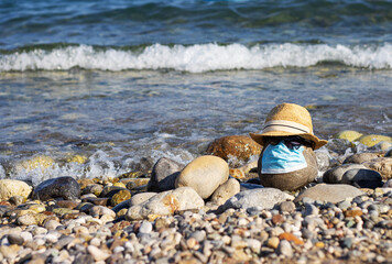 a large stone on the beach with a medical mask from coronavirus Covid-19, a hat and glasses on the background of the sea