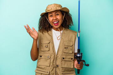 Young african american fisherwoman holding a rod isolated on blue background  receiving a pleasant surprise, excited and raising hands.