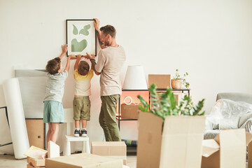 Full length portrait of happy father with two sons hanging pictures on wall while moving in to new home, copy space - obrazy, fototapety, plakaty
