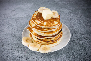 Stack of pancakes on white plate with sliced banana rounds and piece of butter on top, on gray surface