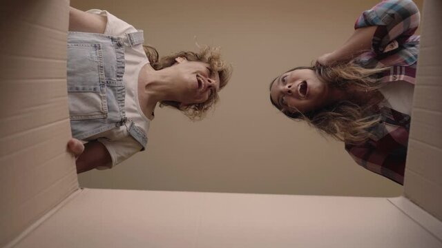 View from inside a cardboard box on two cheerful young Caucasian girls looking into it and smiling. Close-up two smiling girls opening a cardboard box. Woman receiving a package.