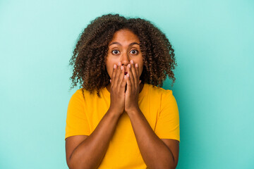 Young african american woman with curly hair isolated on blue background covering mouth with hands looking worried.