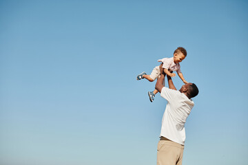 Minimal portrait of happy African-American father playing with cute baby and throwing him against blue sky, copy space - Powered by Adobe