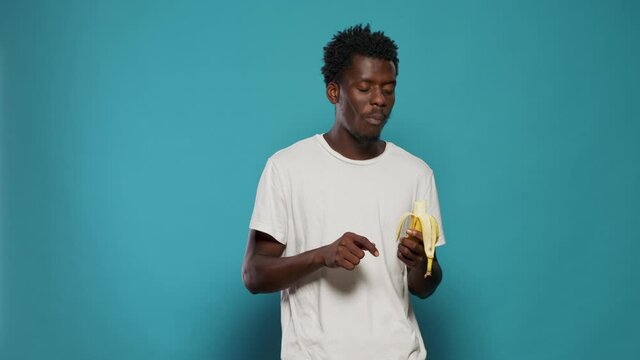 Portrait of casual adult eating banana on camera in studio. Young person enjoying yellow fruit with vitamins for healthy nutrition and fresh diet. Vegetarian man tasting natural snack