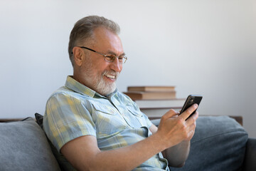 Relaxed joyful middle aged old man in eyeglasses using cellphone software application resting on...