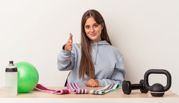 Young caucasian woman sitting at a table with sport equipment isolated on white background stretching hand at camera in greeting gesture.